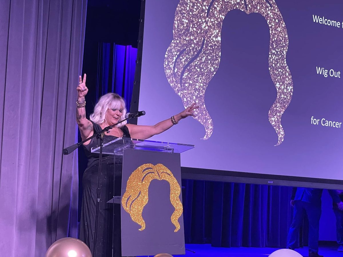 Dana McCool speaking at the Wig Out for Cancer charity event, arms raised in celebration